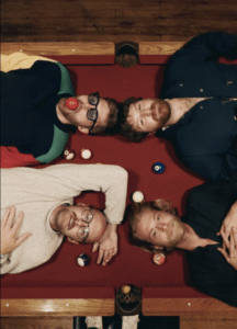 Four men lie on a red pool table surrounded by billiard balls, looking up at the camera.